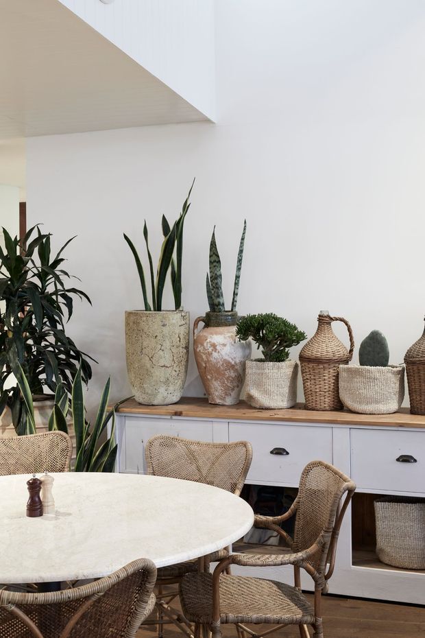 A bright, softly styled dining area with a round pale stone tabletop and woven rattan dining chairs. A white built-in sideboard with timber top occupies the wall, decorated with potted snake plants, greenery, terracotta vessels, woven baskets and natural-textured accessories. White walls and a pale timber ceiling create an airy, organic setting.
