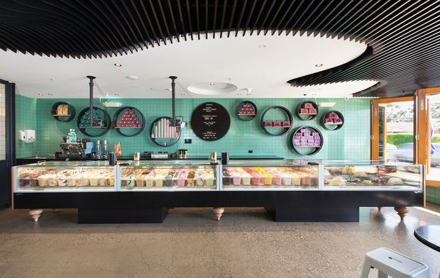 A brightly lit commercial dessert or food shop interior with long refrigerated glass display counters, a turquoise tiled feature wall, circular wall-mounted displays, and a dramatic black slatted ceiling with a large curved white inset. Timber-framed glazing and a small amount of outdoor scenery are visible at the right. The space has polished concrete flooring and a contemporary, playful fit-out.