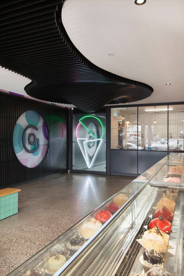 A modern commercial dessert or gelato shop interior with a long glass display counter in the foreground, showcasing colourful desserts. The space features black ribbed wall panels, a sculptural flowing black ceiling installation, polished concrete or terrazzo-like flooring, dark-framed internal glazing, and bright neon geometric signage. A colourful circular graphic feature decorates the rear wall. The overall design is bold, playful, and contemporary.
