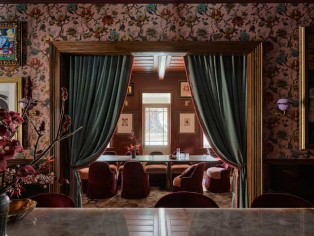 A richly decorated dining or restaurant interior viewed across a polished stone or marble counter. A large timber-trimmed opening with dark draped curtains frames a dining area with a long table, upholstered burgundy chairs, dark wood wall panelling, framed artwork, floral wallpaper, and warm, dramatic lighting. The room has an ornate, theatrical and luxurious character.