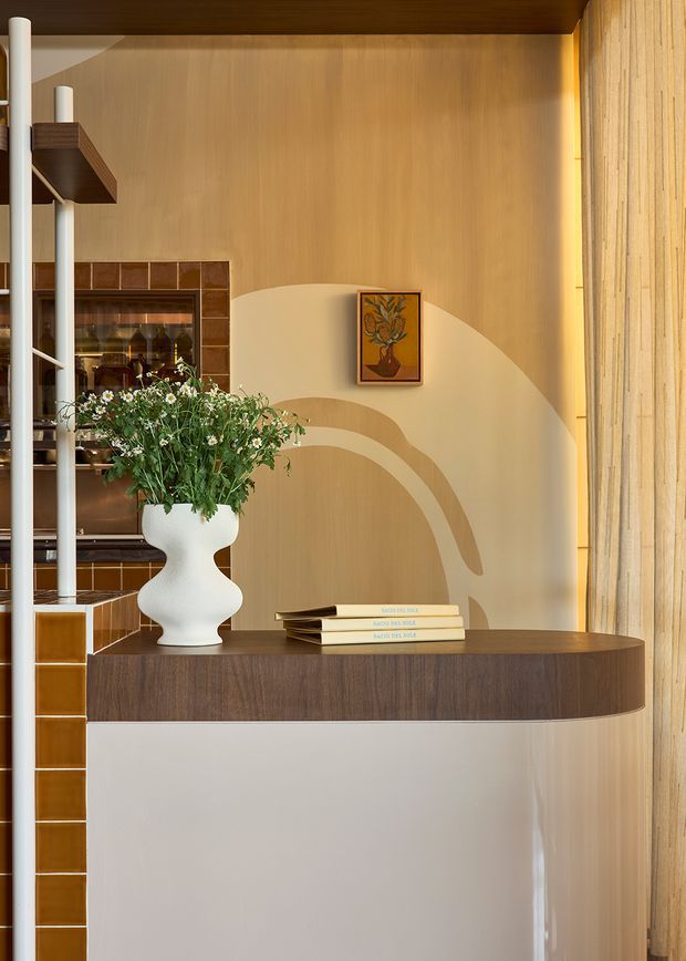 A warm, carefully styled interior vignette featuring a curved built-in console with a dark timber top and glossy light-coloured base. The wall behind it has a soft beige finish with a large abstract curved painted motif, complemented by vertical timber panelling at right. Brown glazed ceramic tiles frame an open shelving or display area on the left, while a sculptural white vase, greenery, books, and a small botanical artwork add decorative detail. Slim white structural or shelving elements and warm concealed lighting contribute to the refined contemporary composition.