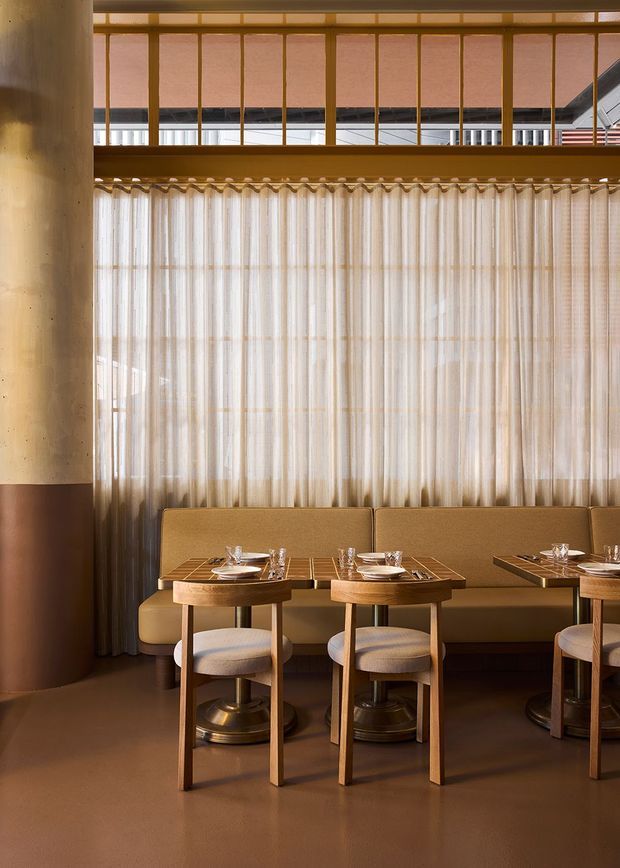 Warmly lit contemporary restaurant dining interior with a row of upholstered banquette seating and small round wood dining tables set with place settings. Sheer floor-to-ceiling curtains diffuse daylight across the rear wall, while a timber-framed mezzanine or upper-level balustrade spans above. A large cylindrical structural column is finished in two-tone warm beige and brown surfaces. The room has polished concrete or resin-like flooring and a refined, softly monochromatic palette of cream, tan, caramel, and warm wood.
