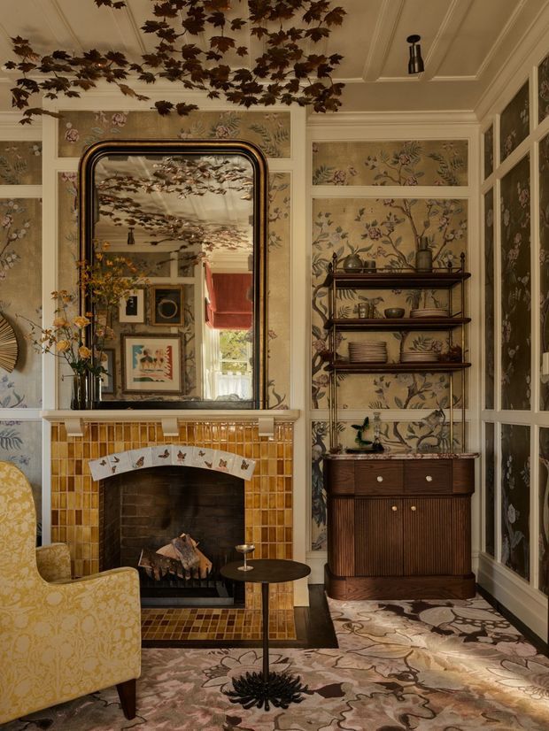 A richly layered traditional sitting room with a dramatic tiled fireplace, ornate floral wallpaper and panelling, a large rounded mirror, patterned carpet, antique-style shelving and cabinetry, and a floral upholstered armchair. Decorative dried foliage is arranged across the coffered ceiling, creating an eclectic, theatrical and warm interior.