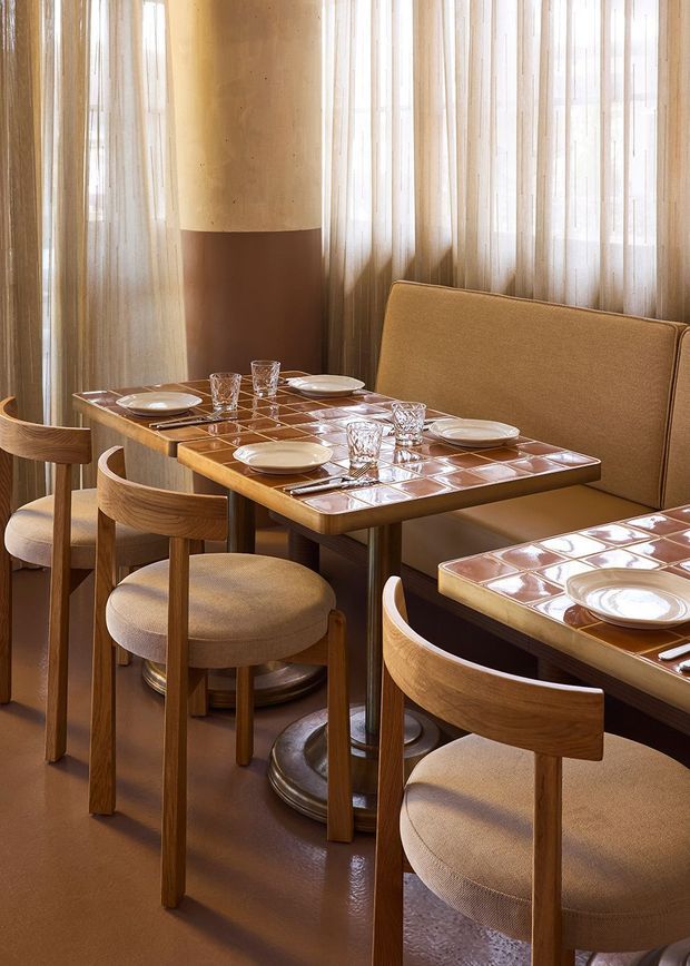 A warmly lit restaurant dining area with compact wooden tables, curved upholstered chairs, and a built-in banquette. The tables feature glossy terracotta-coloured tiled surfaces with place settings and glassware. Sheer curtains filter daylight around a rounded column, creating a refined, intimate hospitality interior.