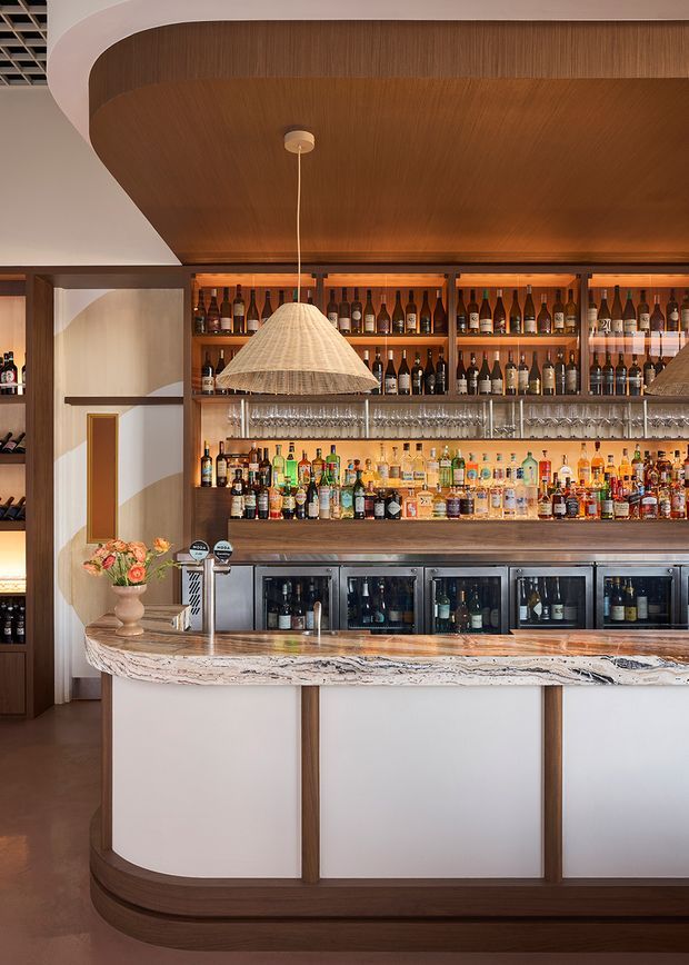 A warmly lit, refined bar interior with a curved timber-clad ceiling and matching dark timber joinery. The built-in back bar features illuminated shelves densely stocked with wine and spirits, glassware, under-counter beverage refrigerators, and a pale veined stone bar top with a curved front. A woven pendant light hangs above the counter, while a floral arrangement and beer taps add detail. The palette is rich in mid-to-dark timber, warm amber lighting, cream surfaces, and natural stone.