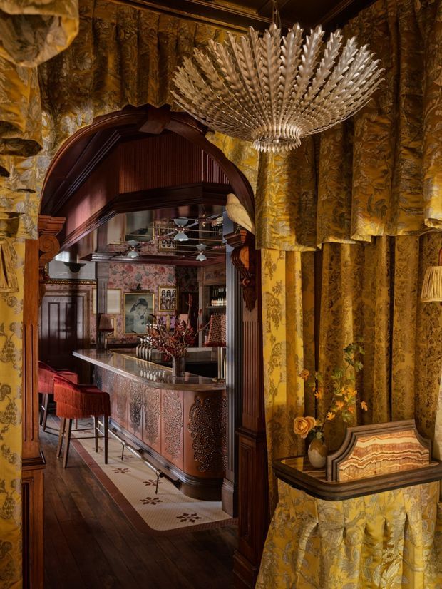 A richly layered, highly decorative interior showing a dramatic home bar beyond an ornate timber arched opening. The bar features a patterned metal-clad counter, red upholstered stools, dark timber joinery, framed artwork, and reflective shelving. The foreground is wrapped in gold and ochre patterned wall and drapery-like finishes, with a large sculptural feathered chandelier overhead and a small built-in display shelf. The atmosphere is theatrical, warm, and opulent, with strong Art Deco-inspired detailing and maximalist styling.