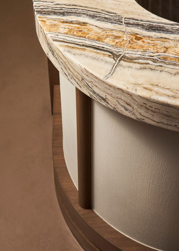 Close-up architectural detail of a curved reception or bar counter with a thick, highly veined natural stone top in cream, tan, grey, and brown tones. The counter has a rounded white textured base, dark timber vertical trim, and a dark timber plinth, creating a refined, sculptural interior feature.