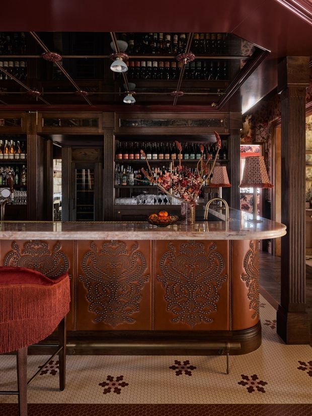A richly detailed, dark-toned bar interior with a curved copper-clad front featuring ornate embossed floral panels, a pale stone or marble countertop, dark timber cabinetry, mirrored bottle shelving, patterned pendant lamps, and decorative mosaic tile flooring. The space has an opulent, intimate atmosphere with strong Art Deco-inspired detailing and warm copper, burgundy, and timber tones.