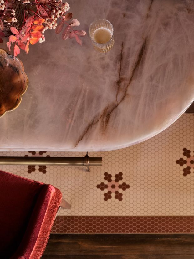 Top-down view of a richly styled bathroom featuring a freestanding oval bathtub with a translucent veined stone or marble-like finish, a wall-mounted bronze-toned tub filler, patterned cream hexagonal tile flooring with burgundy geometric accents, dark timber flooring or trim, and a deep red upholstered chair. Warm autumnal floral styling and a small drink on the bath create a luxurious, moody atmosphere.