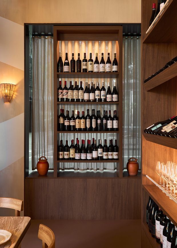 A warm, richly detailed wine storage and display area with a floor-to-ceiling timber-framed glazed wine cabinet holding multiple rows of bottles. Dark timber shelving and cabinetry continue along the right wall, with wine glasses displayed below and additional bottles on angled racks. Sheer curtains, integrated warm lighting, ceramic vessels, and partial timber seating create an intimate hospitality-inspired interior.