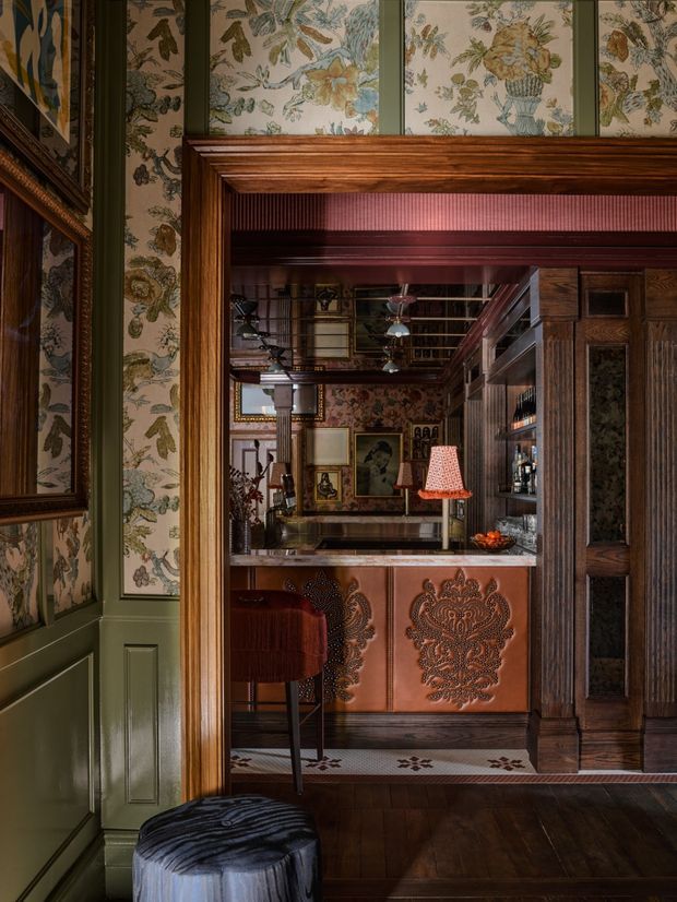A richly layered, moody interior view through a dark timber-framed opening into a compact home bar. The space features botanical wallpaper, deep green painted wall panelling, ornate dark wood cabinetry and shelving, a patterned copper-toned bar front, marble-look countertop, decorative artwork, and warm accent lighting. A reflective surface behind the bar adds depth, while a velvet bar stool and upholstered ottoman sit in the foreground. The overall character is highly decorative, vintage-inspired, and luxurious.