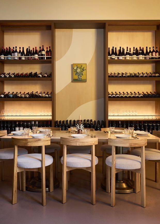 A warmly lit wine-focused dining room or restaurant interior with extensive built-in wooden wine shelving, illuminated display niches, rows of bottles, and glassware. Three compact round timber dining tables are arranged in front, each set with plates, glasses, and cutlery, accompanied by upholstered wooden chairs. The central wall features a pale abstract mural-like finish and a small framed botanical artwork. The atmosphere is refined, intimate, and sophisticated.