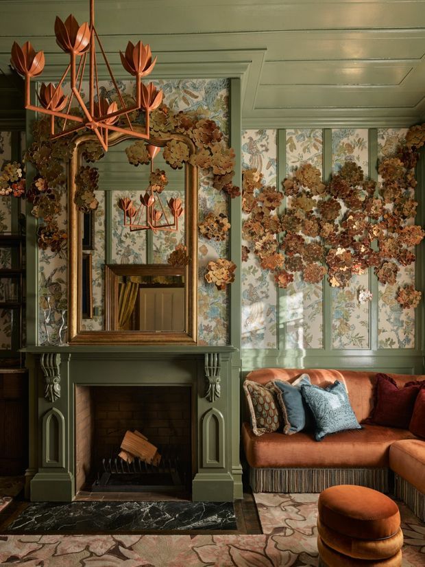 A richly decorated living room centered on an ornate dark green fireplace with carved pilasters, a large framed mirror, floral wallpaper panels, and a sculptural botanical chandelier. A rust-orange upholstered sofa with patterned cushions and a matching rounded ottoman occupies the right side. The room has dramatic layered pattern, warm jewel tones, and traditional detailing, with a dark marble hearth and decorative rug.