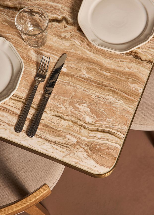 Top-down close-up of a dining table with a highly figured tan and cream stone surface, rounded edge, and warm metallic trim. Cream scalloped-edge plates, clear drinking glass, and dark cutlery are arranged on the table, with upholstered dining chairs partially visible around it. The composition is warm, refined, and material-focused.