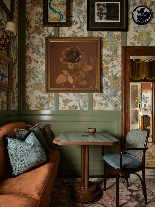 A richly layered traditional sitting and dining nook with a warm caramel leather banquette, patterned cushions, a small dark wood pedestal table, and a vintage upholstered chair. The walls feature green-painted wainscoting and ornate botanical wallpaper, densely decorated with framed artwork and a decorative plate. A mirror-framed doorway reveals an adjoining room, while a patterned rug and deep, moody lighting reinforce the intimate, collected atmosphere.