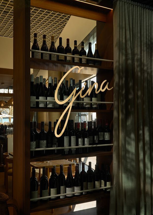 A vertically framed interior detail of a wine display shelving unit in a dimly lit hospitality space. Multiple rows of wine bottles are arranged on dark timber shelves with slim metal rails, behind a large illuminated cursive “Gmina” sign. A ribbed translucent glass or textured vertical screen occupies the right side, while a decorative gridded ceiling and warm timber finishes are visible in the background. The atmosphere is warm, intimate, and refined.