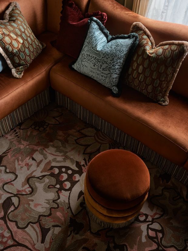 Close-up interior view of a richly layered sitting area featuring a rust-orange upholstered sectional sofa with decorative fringe, patterned throw pillows, an ornate floral area rug, and a round stacked upholstered ottoman. Warm low lighting and deep earthy tones create an eclectic, cozy atmosphere.