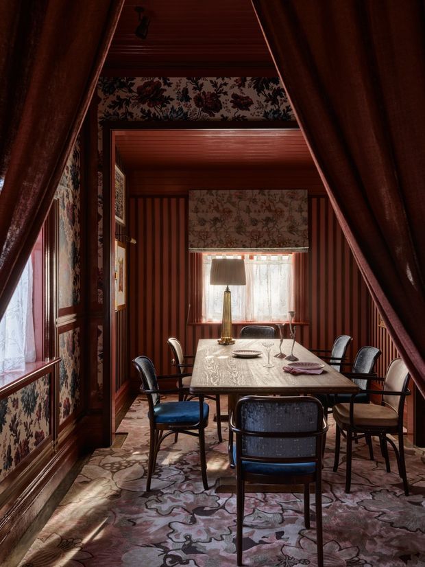 A richly layered dining room viewed through dark timber-framed openings, featuring a rectangular dining table surrounded by upholstered chairs, floral patterned carpet and wallpaper, deep red vertical wall panelling, timber ceilings and trim, and curtained windows. The low warm lighting and ornate botanical patterns create a dramatic, intimate, traditional atmosphere.