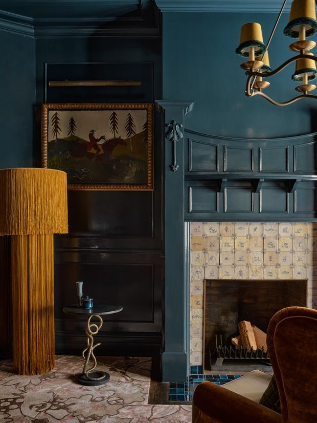 A richly atmospheric, dark-toned traditional sitting room focused on a fireplace wall. Deep blue-black painted timber wall panelling surrounds a tiled fireplace with patterned cream tiles and a dark timber mantel area. A vintage hunting landscape artwork, sculptural side table, fringed floor lamp, brass chandelier, plush brown armchair, and patterned rug create a layered, luxurious interior.