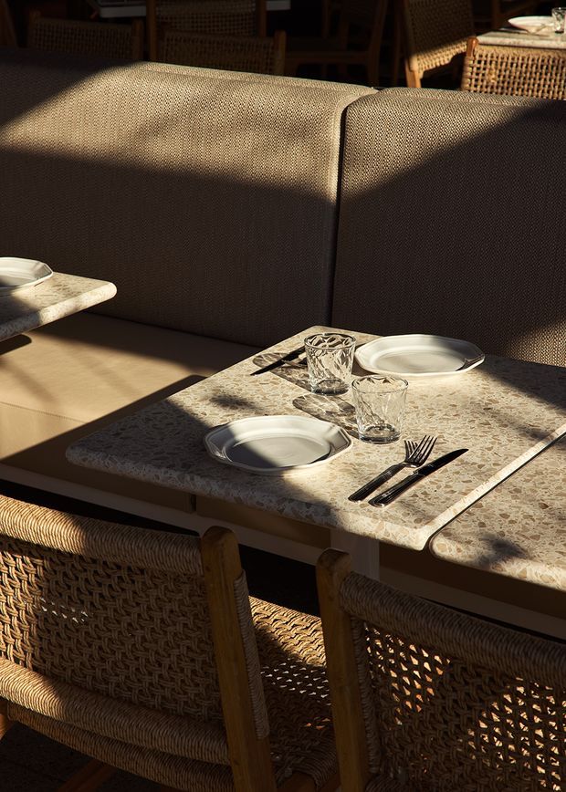 Close-up of a warmly lit restaurant dining setting with a built-in upholstered banquette, woven rattan dining chairs, and a rectangular table topped with a mottled stone-look surface. Place settings include ceramic plates, faceted drinking glasses, and dark cutlery. Strong sunlight and shadows create a relaxed, intimate atmosphere.