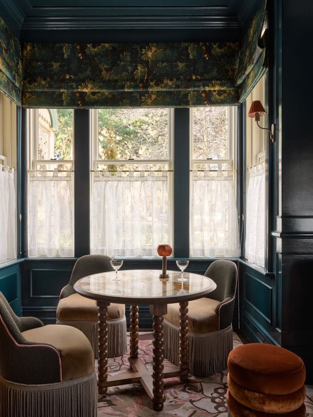 A richly styled, compact dining nook with dark teal-blue wall panelling, a moulded coffered ceiling, and tall windows dressed with botanical Roman blinds and sheer curtains. A round marble-topped pedestal table is surrounded by upholstered fringe-trimmed armchairs, with a small central lamp and glassware. The room has a moody, elegant, highly layered decorative character.