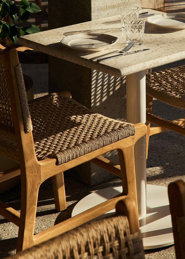 Close-up of a warmly lit dining setting with a cream stone-look rectangular tabletop, rounded light timber legs, and woven natural-fibre dining chairs. The table is set with ceramic plates, cutlery, and textured drinking glasses. Strong sunlight casts patterned shadows across the chairs and floor, creating a relaxed, natural atmosphere.