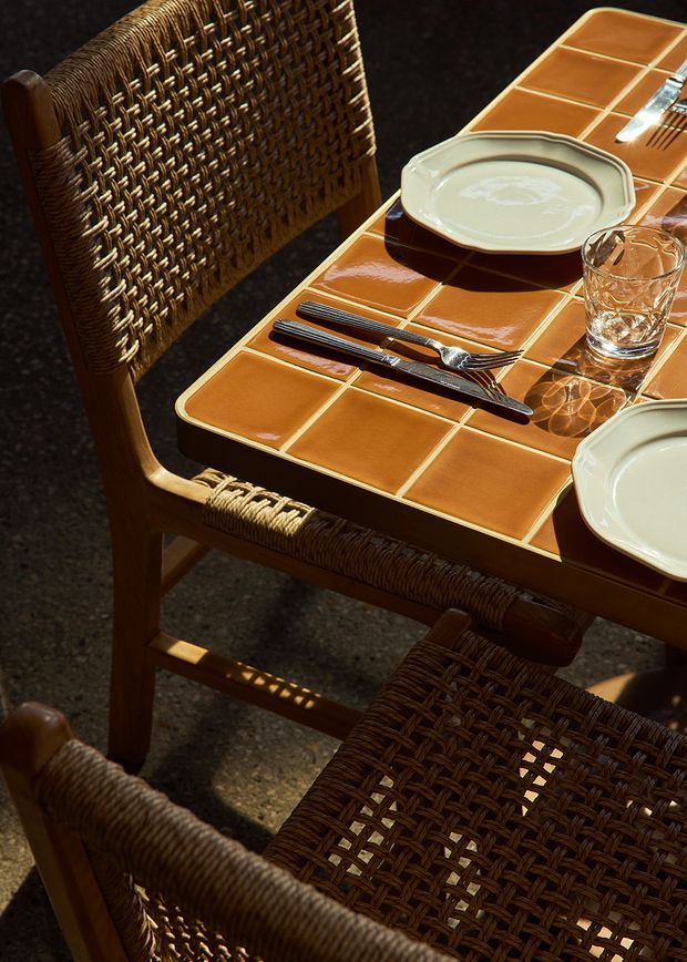 Close-up view of a warmly lit dining setting with a rectangular terracotta-orange tiled tabletop, cream ceramic plates, clear drinking glasses, cutlery, and woven cane dining chairs with wooden frames. Strong sunlight creates dramatic shadows across the table and chairs, giving the scene a rustic, tactile character.