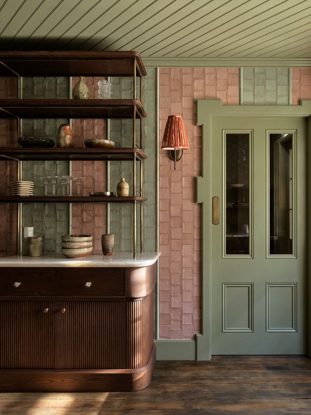 A richly detailed interior vignette featuring a dark fluted timber built-in cabinet and open shelving, displaying ceramics and glassware. The cabinet has a pale stone or composite benchtop. Behind it, small-format glazed tiles in muted pink and sage green cover the wall. A sage green paneled door with narrow glazed inserts and decorative stepped trim occupies the right side. The ceiling is clad in narrow painted timber boards, and the dark timber floor, warm lighting, and vintage furnishings create a refined, moody atmosphere.