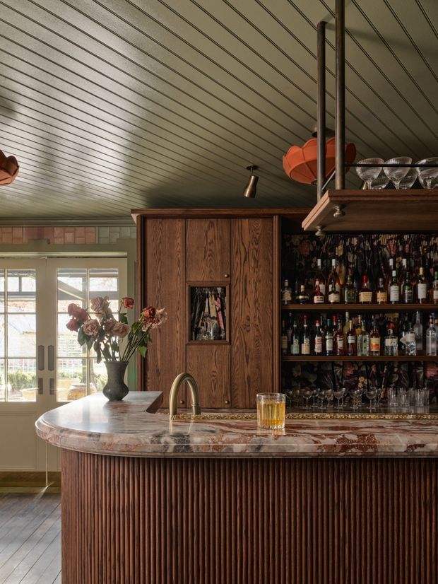 A richly detailed residential home bar with a curved island featuring a heavily veined stone benchtop and vertically fluted dark timber front. Behind it is extensive dark timber cabinetry with open shelving stocked with bottles and glassware, plus a concealed bar cabinet. The room has a pale green beadboard-style ceiling, warm timber flooring, French doors, vintage-style lighting, and layered traditional decorative details.
