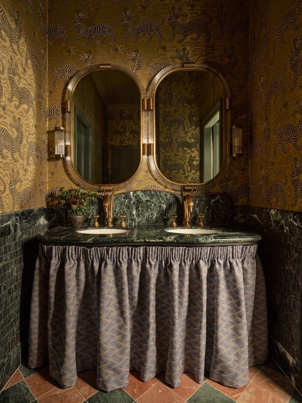 A richly decorated, compact double-vanity bathroom with two oval inset mirrors in ornate metallic frames, dark green marble vanity top and splashback, patterned brown-gold wallpaper, dark wall tiling, brass fixtures, and a pleated fabric vanity skirt. The room has an opulent, theatrical character with warm low lighting and patterned terracotta and green floor tiles.