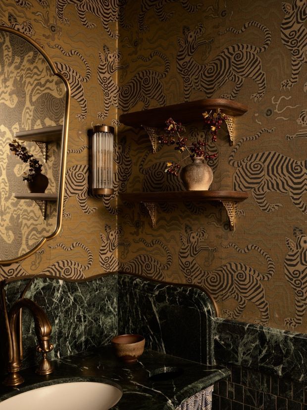 A richly styled, dark and luxurious bathroom vanity vignette. The space features dramatic patterned wallpaper with animal and botanical motifs, dark green veined marble across the benchtop and backsplash, a curved integrated basin, aged brass tapware, a tall ribbed wall light, an organic ceramic vessel, and dark timber display shelves with ornate brass brackets. A large rounded-edge mirror is partially visible at left.