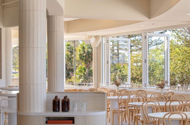 Bright coastal-inspired dining area with extensive openable glazing, curved tiled columns, a curved tiled service counter, and numerous light timber bentwood chairs and round tables. Lush greenery is visible outside, while the interior uses soft cream and white finishes with warm natural timber accents.