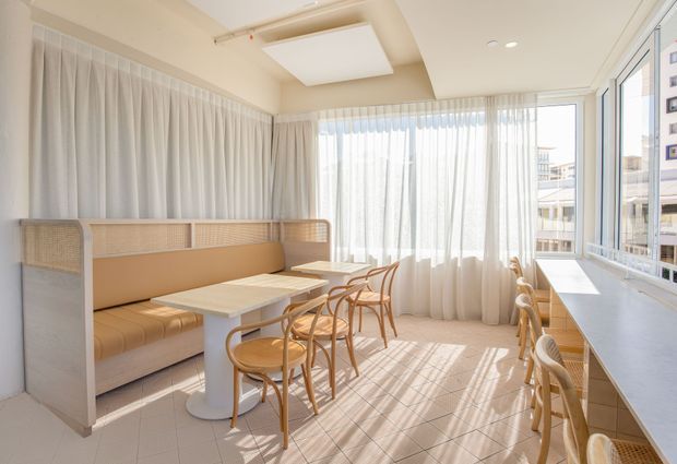 Bright, compact dining and breakfast area with a built-in upholstered banquette, cane-panel dividers, light timber dining chairs, and a long white counter with stools. Full-height sheer curtains and expansive corner glazing fill the room with natural light, while a large rectangular ceiling light and pale tiled floor create a clean, airy contemporary setting.