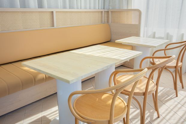 A bright, warm-toned dining area with a built-in tan upholstered banquette, pale timber tables, and curved natural timber cane-back chairs. Soft daylight filters through sheer curtains, creating a calm hospitality-style interior with subtle woven textures and rounded detailing.