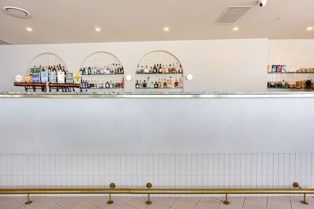 A bright, refined commercial bar interior viewed from the customer side. A long pale bar counter spans the foreground, with a brass-toned foot rail and tiled floor below. Behind it, a white wall incorporates three large arched recessed bottle displays and additional shelving, while recessed ceiling lights and ventilation provide even illumination.