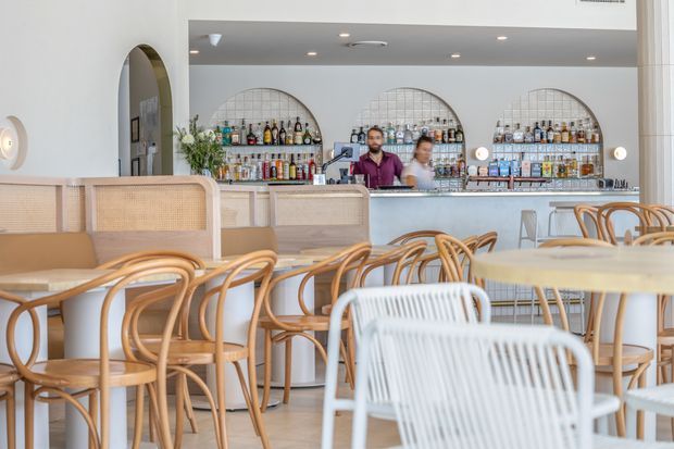 Bright contemporary hospitality interior showing a café or restaurant dining area with light timber and cane chairs, pale upholstered banquette seating, a long service bar, and an open back bar displaying bottles and glassware. Arched wall openings and shelving add a soft architectural feature, with warm timber accents against white walls and ceilings.