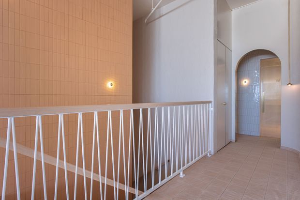 A bright upper-level hallway or landing with a white geometric metal balustrade overlooking a stairwell. The space features pale terracotta-pink tiled wall cladding, light beige tiled flooring, white walls, warm wall sconces, and an arched opening leading to a tiled area beyond.