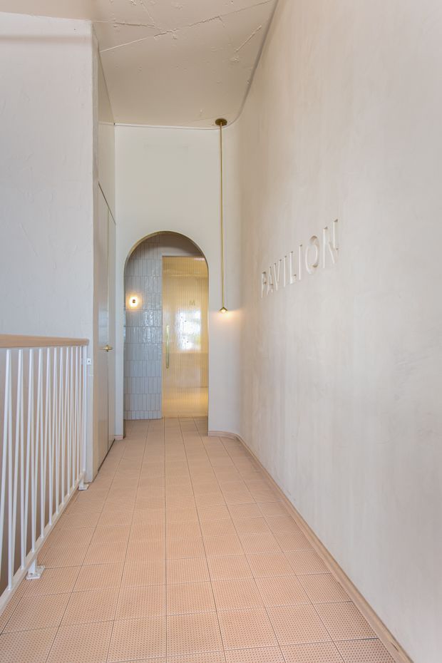 A narrow, light-filled hallway with pale tiled flooring and textured off-white walls. A white balustrade lines the left side, while a minimalist pendant light hangs beside raised wall lettering. At the end, an arched opening leads to a tiled bathroom or shower area with a translucent golden glass door.