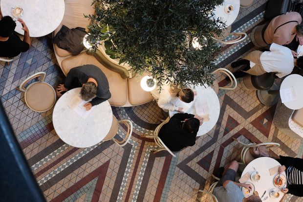 Overhead view of a stylish café or restaurant interior with multiple small round marble-topped tables, curved upholstered banquette seating, bentwood-style chairs, decorative hanging greenery and globe lighting. The floor features an elaborate geometric patterned tile mosaic in muted terracotta, blue, cream and grey tones, with several patrons seated at the tables.