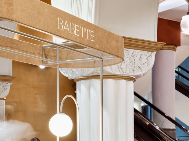 Close-up view of an ornate retail or commercial interior featuring a prominent Babette-branded display structure, sculptural white fluted columns, elaborate classical plaster ornamentation with gold-toned mouldings, and a dark stairway in the background. The space combines historic decorative architectural detailing with contemporary retail display elements and lighting.