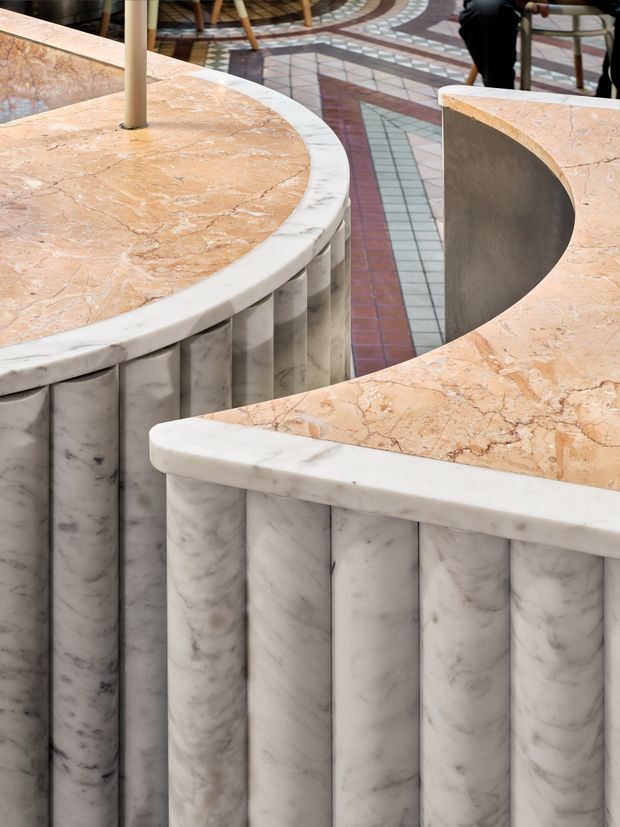 Close-up architectural detail of curved counters or built-in furniture clad in veined marble. The foreground features white-grey marble fluted vertical panels with rounded white marble edge caps and warm peach-beige marble benchtops. A patterned geometric tiled floor and partial seating are visible in the background, suggesting a refined interior commercial or hospitality setting.