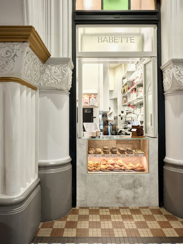 A small bakery or café service counter set within an ornate historic interior. The framed service opening has black trim and frosted glass signage reading “BABETTE,” with a marble-look counter and illuminated pastry display filled with breads and pastries. Behind it is a compact commercial kitchen with open shelving and equipment. The surrounding space features decorative white carved columns, classical mouldings, dark grey lower walls, and a patterned small-format tiled floor.