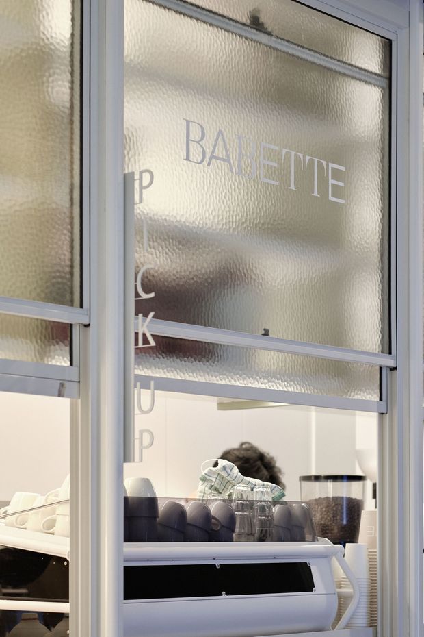 Close-up view through a storefront window with frosted textured glazing and white-painted metal framing. The glass bears partial lettering, including “BARETTE,” while a commercial coffee counter with an espresso machine, cups, glassware, and a person is visible inside.