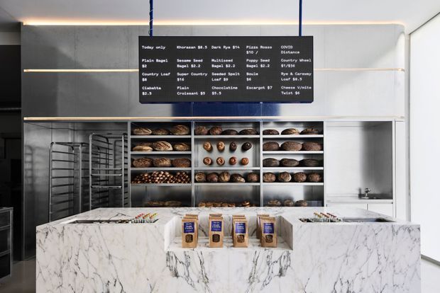 Contemporary bakery retail interior with a prominent white marble-patterned service counter in the foreground, stainless-steel bread display shelving and baking racks behind it, and a dark overhead menu board. The space has clean white walls, dark grey panels, integrated linear lighting, and a bright, refined commercial fit-out.