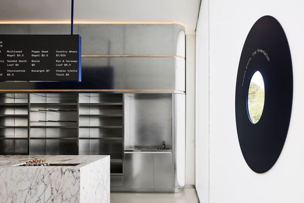 A refined contemporary commercial food-service interior, likely a café or dessert bar. The space features a large white marble island in the foreground, extensive brushed stainless-steel shelving and cabinetry, a dark menu board, integrated warm linear lighting, and a distinctive black oval wall feature with a circular window or opening. The composition is clean, geometric, and minimalist with a restrained monochrome palette.