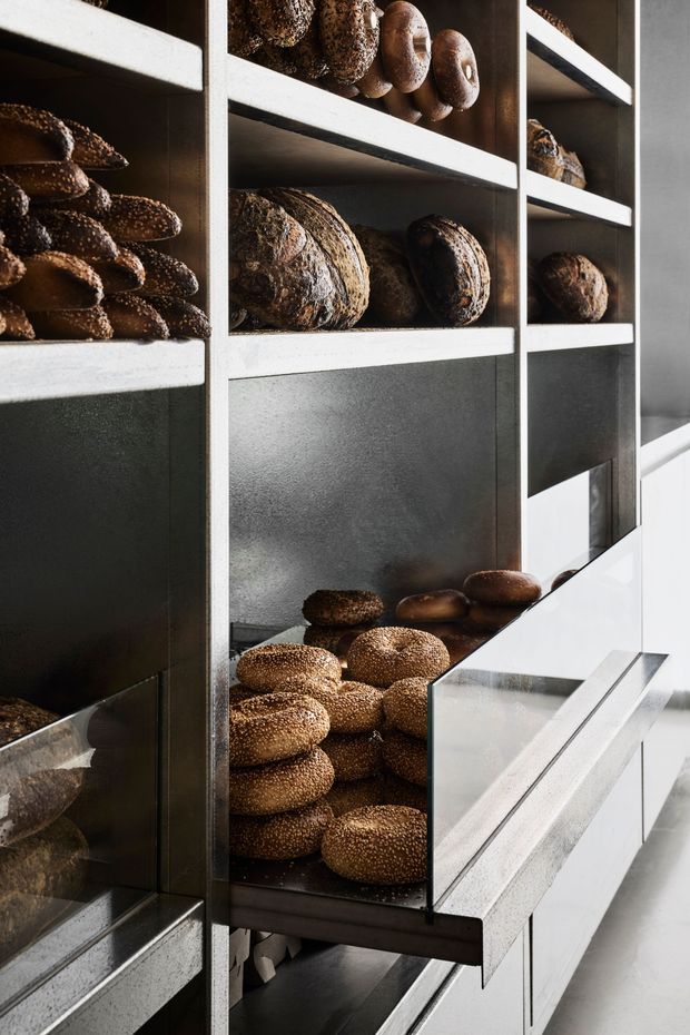 Close-up view of a contemporary bakery or bread-display interior, with dark metal-framed shelving and glass-fronted compartments filled with loaves, rolls, and seeded bread. Strong natural light enters from the right, highlighting the contrast between the dark backing surfaces, pale shelving, and stainless-steel or light-toned counter elements.