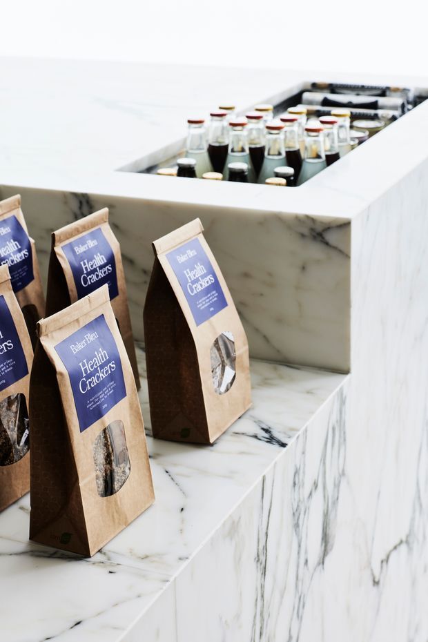 Close-up of a refined retail or hospitality counter display featuring white marble with prominent dark veining. Packaged crackers are arranged on the front shelving, while bottled beverages sit in a recessed ice or display compartment on the countertop. The image focuses on the stone joinery and product presentation, with a bright, clean, high-end aesthetic.