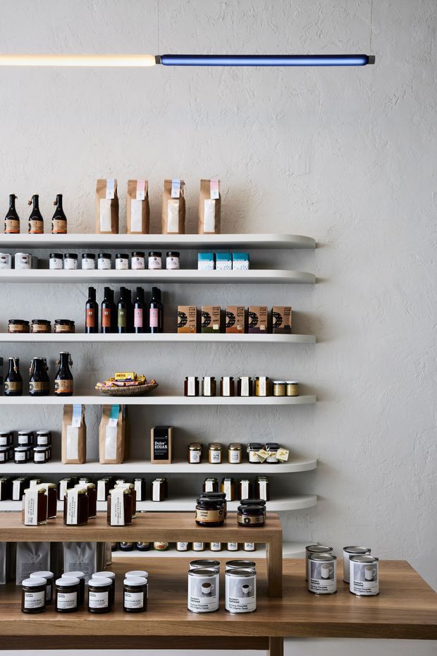 A neatly arranged retail shelving display against a textured light-grey plaster wall. Multiple floating shelves and a timber display counter hold jars, bottles, tins, boxed goods, and paper-wrapped products. A slim linear wall light runs horizontally above the shelves. The composition is minimal and product-focused, resembling a boutique shop or commercial showroom interior.