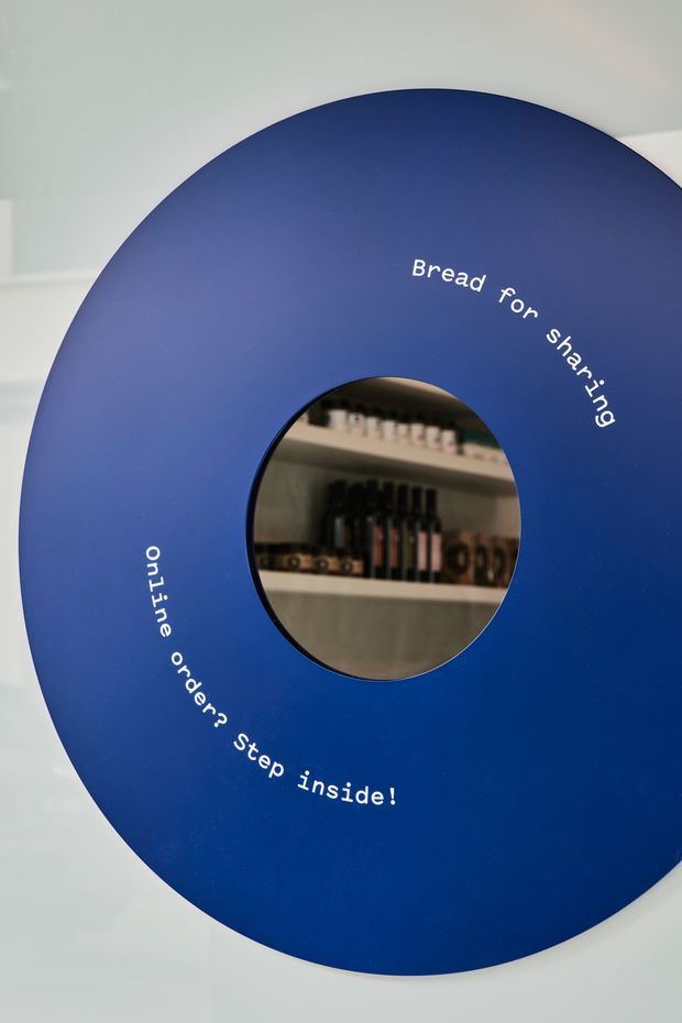 Close-up of a large circular blue wall sign or display panel with a central round opening revealing shelves of bottles behind it. White curved text reads “Bread for sharing” and “online orders? step inside!”. The image appears to show a retail or hospitality venue entrance feature with a playful, graphic design.