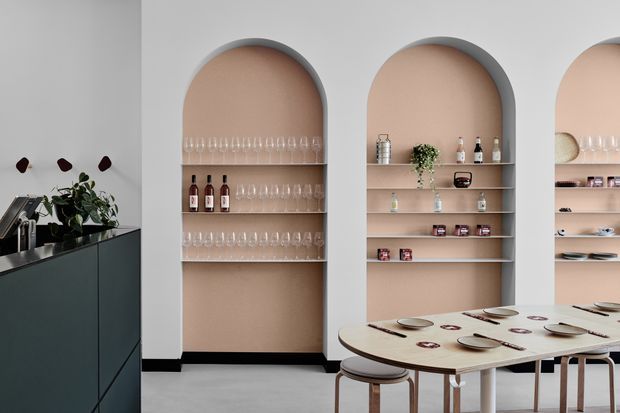 Contemporary dining and entertaining space with a pale grey wall, three tall arched recessed display niches in warm beige, and open shelving holding glassware, bottles, ceramics and small decorative objects. A dark green-black kitchen or bar counter with sink occupies the left foreground, while a light timber dining table with place settings and stools fills the right side. The scheme is restrained and refined with strong arched joinery details and soft neutral tones.
